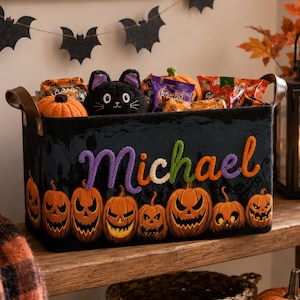 May include: A black felt storage basket with brown leather handles, featuring the name Michael embroidered in purple and orange. The basket is decorated with a row of orange jack-o'-lanterns and contains various small plush toys and candy bags.