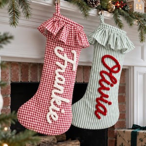 Puede incluir: Dos calcetines navideños con puños de volantes. El calcetín izquierdo es de cuadros vichy rojos y blancos con el nombre Frankie en letras blancas. El derecho es de rayas verdes y blancas con el nombre Olivia en letras rojas. Ambos tienen lazos.