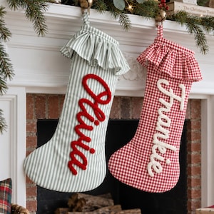 Puede incluir: Dos calcetines de Navidad personalizados colgados de una repisa. El calcetín izquierdo es de rayas verde claro y blanco con un puño con volantes y el nombre Olivia en rojo. El derecho es de cuadros vichy rojo y blanco con el nombre Frankie en blanco.
