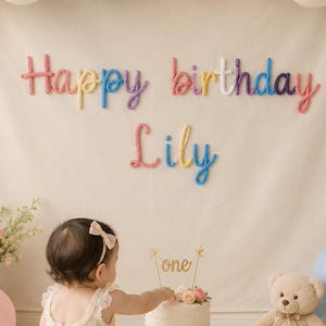 Op de afbeelding: Een crèmekleurige stoffen achtergrond met de tekst Happy birthday Lily in meerkleurige garenletters. De decoratie bestaat uit pastelkleurige ballonnen, een kleine taart met een gouden one-topper en een beige pluche beer.