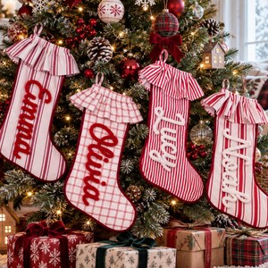 Puede incluir: Cuatro calcetines de Navidad colgados en un árbol, cada uno con un borde con volantes y un nombre personalizado en letra cursiva. Tres calcetines tienen rayas verticales rojas y blancas, mientras que uno tiene un patrón de cuadros. Los nombres son Emma, Olivia, Lucy y Noah.