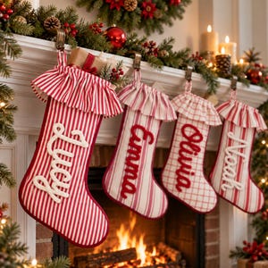 Puede incluir: Cuatro calcetines navideños colgados de una repisa, cada uno con un borde con volantes y un nombre bordado en letra cursiva blanca o roja. Los calcetines muestran varios patrones rojos y blancos, incluyendo rayas y un diseño de cuadros.