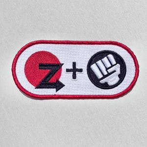 May include: An oval-shaped embroidered patch with a white background and a red border. It features a red circle containing a black letter Z, a black plus sign, and a black circle containing a white fist icon, all rendered in a clean, bold style.