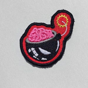 May include: An embroidered patch featuring a black bomb shape with a pink brain inside the top section. The bomb has a white fuse with a yellow starburst at the end, a white curved mouth with teeth, and a red border outlined in black thread.