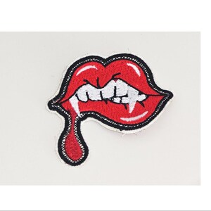 May include: An embroidered patch featuring a pair of bright red lips with white fangs biting the lower lip. A single drop of red liquid drips from the corner of the mouth. The entire design is outlined in thick black thread with white highlights.