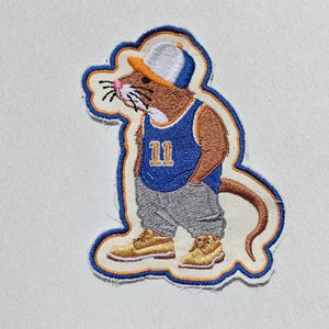 May include: An embroidered patch featuring a brown mouse wearing a white and orange baseball cap, a blue basketball jersey with the number 11 in yellow, gray baggy pants, and tan boots. The patch has a thick blue and orange border outline.