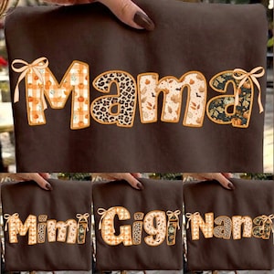 Könnte beinhalten: Ein dunkelbraunes T-Shirt mit dem Wort "mama" in großen, stilisierten Buchstaben. Jeder Buchstabe hat ein herbstliches Muster, darunter orangefarbene Karos mit Kürbissen, Leopardenmuster, Herbstblätter und ein dunkles Blumenmuster.