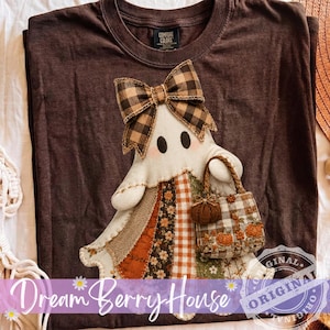 May include: A dark brown t-shirt featuring a central applique of a ghost wearing a brown and black buffalo plaid bow. The ghost has a patchwork-style dress with floral and gingham patterns and carries a small tote bag with pumpkin embroidery.