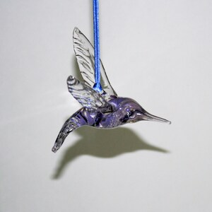 Puede incluir: Un adorno de colibr&iacute; de vidrio morado con alas transparentes que cuelga de una cinta azul.