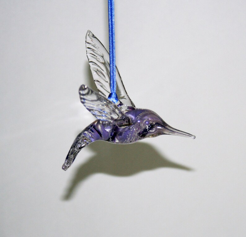 Puede incluir: Un adorno de colibr&iacute; de vidrio morado con alas transparentes que cuelga de una cinta azul.