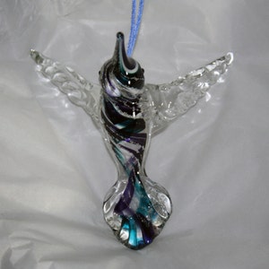 Puede incluir: Un adorno de colibr&iacute; de vidrio con cuerpo y alas transparentes, y una cola colorida con remolinos de morado, azul verdoso, negro y blanco.