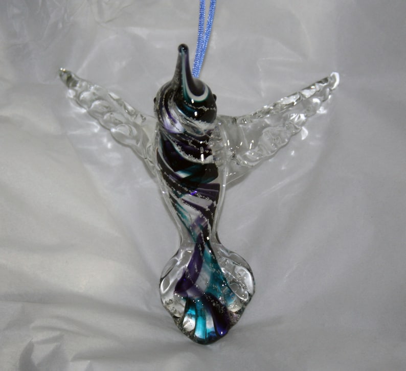 Puede incluir: Un adorno de colibr&iacute; de vidrio con cuerpo y alas transparentes, y una cola colorida con remolinos de morado, azul verdoso, negro y blanco.