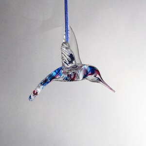 Puede incluir: Un adorno de colibr&iacute; de vidrio con detalles azules, rojos y blancos. El adorno est&aacute; colgado de una cuerda azul.