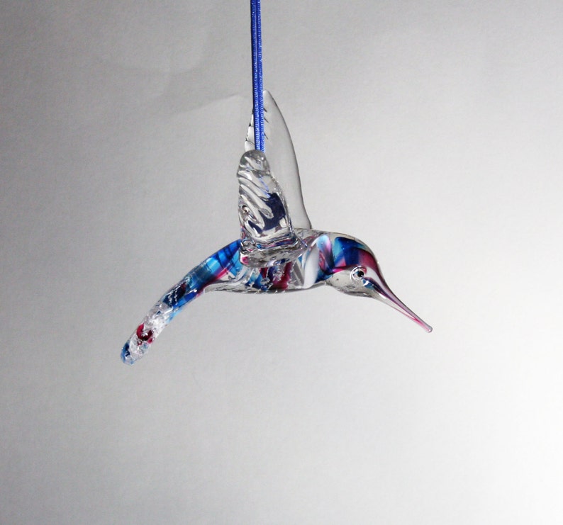 Puede incluir: Un adorno de colibr&iacute; de vidrio con detalles azules, rojos y blancos. El adorno est&aacute; colgado de una cuerda azul.