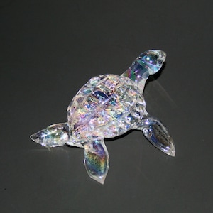 May include: A clear glass turtle figurine with iridescent detailing. The turtle is facing right and has a detailed shell.