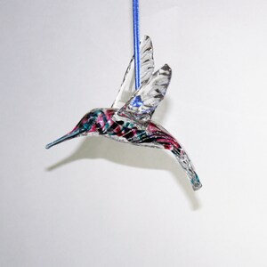 Puede incluir: Un adorno de colibr&iacute; de vidrio transparente con detalles pintados en rosa, azul y negro. El adorno est&aacute; colgado de una cuerda azul.