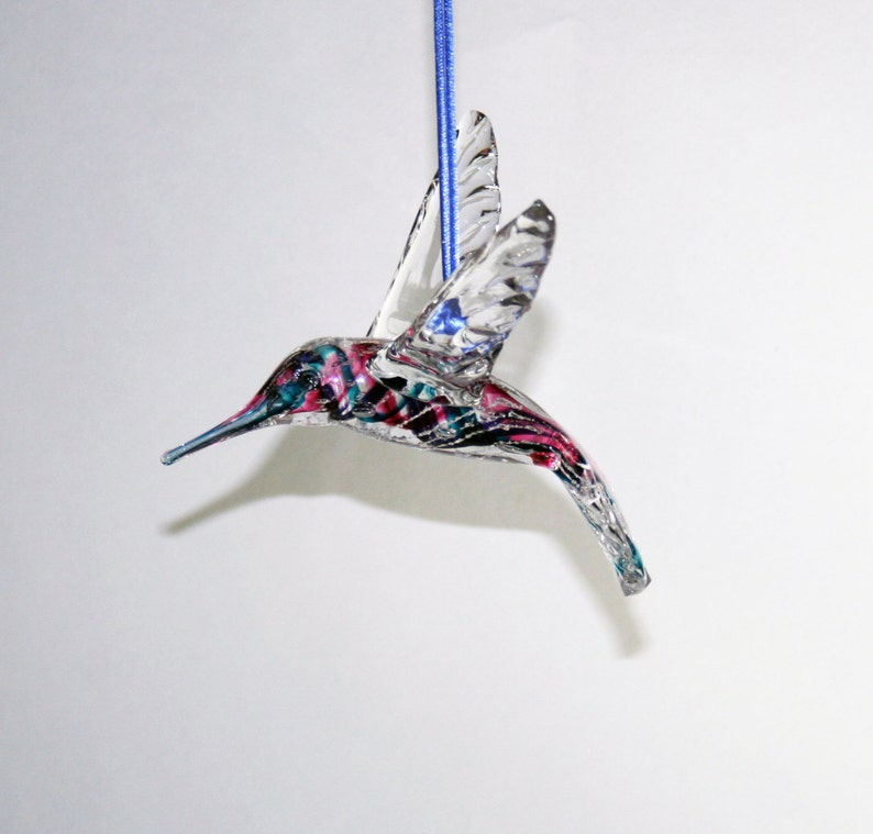 Puede incluir: Un adorno de colibr&iacute; de vidrio transparente con detalles pintados en rosa, azul y negro. El adorno est&aacute; colgado de una cuerda azul.