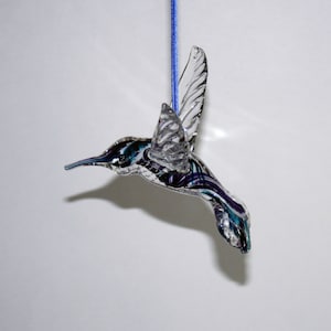 Puede incluir: Un adorno de colibr&iacute; de vidrio con alas transparentes y un cuerpo morado y azul. El adorno est&aacute; colgado de una cuerda azul.