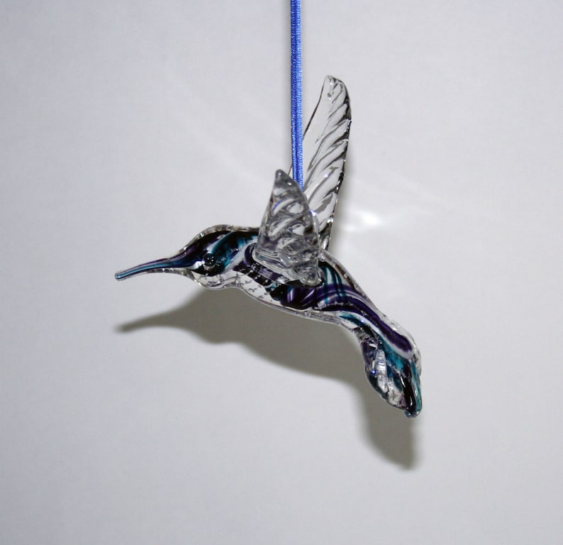 Puede incluir: Un adorno de colibr&iacute; de vidrio con alas transparentes y un cuerpo morado y azul. El adorno est&aacute; colgado de una cuerda azul.