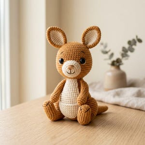 May include: A brown and cream-coloured crocheted kangaroo stuffed animal. The toy features large upright ears, dark plastic eyes, a stitched nose and mouth, and a prominent cream-coloured belly patch. The kangaroo is seated in an upright position.