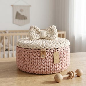 May include: A round, hand-crocheted storage basket with a dusty pink base and a cream-colored lid topped with a large, matching bow. A small tan leather tab with a heart cutout and a rectangular paper tag with text are attached to the front.
