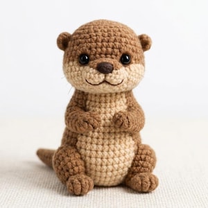 May include: A small, handcrafted crochet otter toy featuring a two-tone design with dark brown yarn for the head, back, and limbs, and a lighter beige yarn for the face, chest, and belly. It has two black plastic safety eyes and a dark nose.