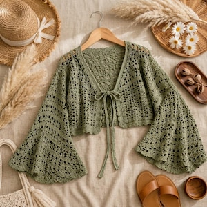 May include: A sage green crochet bolero cardigan featuring long, flared bell sleeves, an open front design with a delicate tie closure, and a scalloped hemline. The garment is crafted with an intricate, open-work knit pattern throughout.