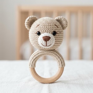 May include: A crochet bear rattle featuring a beige yarn head with a white muzzle, dark brown embroidered nose and mouth, and black safety eyes. The head is attached to a smooth, circular light-colored wooden ring handle for easy grasping.