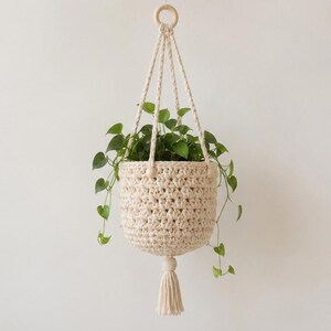 May include: A cream-colored crocheted hanging plant basket featuring a thick, textured stitch pattern. The basket is suspended by matching crocheted cords attached to a circular wooden ring at the top and finished with a decorative tassel at the bottom.