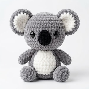 May include: A plush koala toy made of gray and white yarn. The koala features a large, round head with prominent, fluffy white-centered ears, a black nose, and dark, glossy eyes. It has a white belly patch and a soft, textured crochet finish.