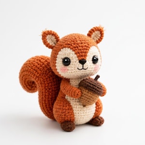 Puede incluir: Una pequeña ardilla de ganchillo hecha a mano con hilo naranja y crema. La ardilla tiene grandes ojos de seguridad negros, mejillas rosadas bordadas y una cola tupida. Sostiene una pequeña bellota de ganchillo marrón en sus patas delanteras.