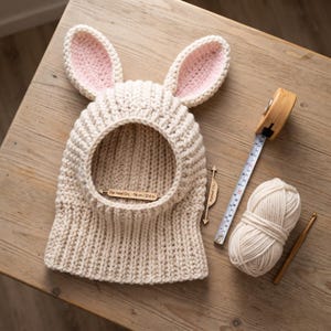 May include: A cream-colored crocheted balaclava with two rabbit ears featuring pink inner accents. A wooden measuring tool inside the opening indicates a flat hood circumference of 58 cm or 22.8 inches. Includes a ball of yarn and a crochet hook.