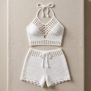 Puede incluir: Un conjunto de dos piezas de ganchillo blanco que consta de un top corto estilo halter y pantalones cortos a juego. El top presenta un escote con patrón de rejilla y cierre de lazo. Los pantalones cortos incluyen cintura con cordón y dobladillo festoneado.