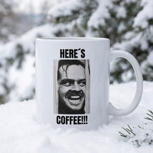 Puede incluir: Una taza de cerámica blanca con un gráfico en blanco y negro de un hombre mirando a través de una puerta rota. El texto sobre la imagen dice HERE'S y el texto debajo de la imagen dice COFFEE!!! en letras mayúsculas negras en el frente.