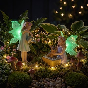 May include: Two fairy figurines with glowing green wings in a garden setting. One fairy stands holding a silver watering can, while the other kneels beside a small dog in a wooden trough. The scene includes moss, small mushrooms, and fairy lights.