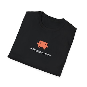 May include: A black t-shirt featuring a small, orange, pixelated character graphic centered above the white text that reads > Human : turn. The shirt is folded neatly, showcasing the crew neckline and the short sleeve construction.