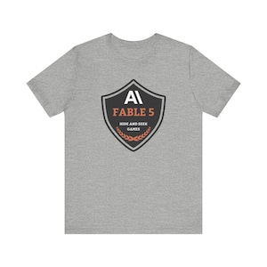 May include: A heather gray short-sleeve t-shirt featuring a black shield-shaped emblem on the chest. The emblem displays the white text AI, the orange text FABLE 5, the white text HIDE AND SEEK GAMES, and an orange laurel wreath graphic at the base.