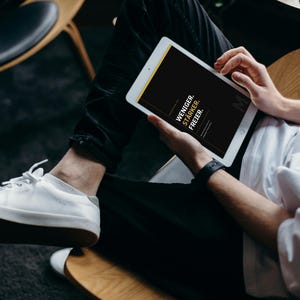 Peut inclure: Une tablette blanche affichant un écran noir avec le texte blanc WENIGER. STÄRKER. FREIER. La tablette est tenue par une personne portant un t-shirt blanc, un pantalon noir et des baskets blanches, assise sur une chaise en bois à coussin noir.