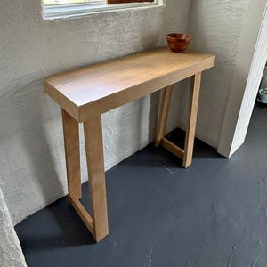 May include: A light brown wooden console table with a rectangular top and four straight legs connected by side supports. A small, round, dark brown wooden bowl sits on the right side of the table surface. The furniture features a smooth finish.