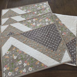 May include: A set of rectangular quilted placemats featuring a geometric chevron pattern. The fabric displays a mix of floral prints in shades of white, orange, and green, combined with solid white and dark brown floral patterned sections with stippled quilting.