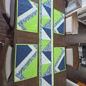 May include: A quilted table runner and four matching rectangular placemats featuring a geometric pattern in lime green, navy blue, and a floral print. The fabric pieces are stitched with a swirling quilting design and finished with lime green binding.