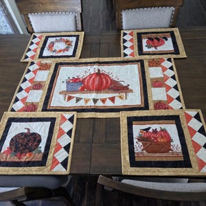May include: A quilted table runner and four matching placemats featuring autumn-themed designs. The fabric displays pumpkins, gourds, and a basket, accented by black, orange, and white diamond-patterned borders. The center piece includes the word harvest.