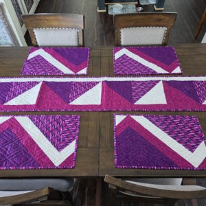 May include: A quilted table runner and four matching rectangular placemats featuring a geometric pattern in shades of purple, magenta, and white. The fabric pieces display a repeating zigzag design with stitched lines and a textured finish.