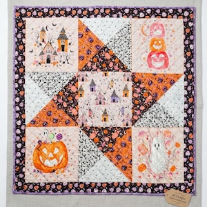May include: A square Halloween-themed quilt featuring a patchwork design of orange, black, and white fabric squares with illustrations of pumpkins, ghosts, and houses. A small brown tag in the corner displays the text BOO-tiful Hand-stitched Quilt.