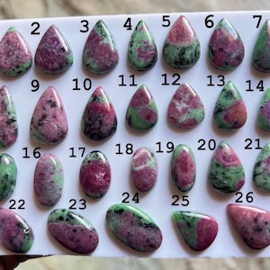 May include: A collection of twenty-six polished ruby in zoisite cabochons arranged on a white display card. Each gemstone features a unique blend of vibrant magenta-pink ruby inclusions contrasted against a mottled green and black zoisite matrix.