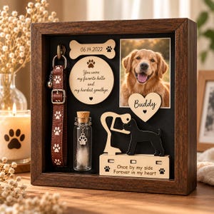 May include: A dark wood shadow box memorial frame containing a brown dog collar with white paw prints, a glass vial with fur, a photo of a golden retriever, and wooden cutouts with the text Buddy, 06.14.2022, and Once by my side Forever in my heart.