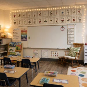 May include: A classroom interior featuring a white board, an alphabet wall display, and a wooden rocking chair with a green patterned cushion. Student desks with name tags, a book rack, and string lights decorate the learning environment.