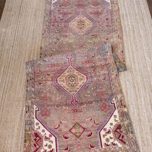 May include: A rectangular area rug featuring a taupe background with a central diamond medallion in cream, accented by intricate geometric patterns in shades of magenta, blue, green, and white. The border displays a repeating linear motif.