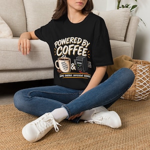 May include: A black short-sleeve t-shirt featuring the text "POWERED BY COFFEE" above a graphic of a coffee mug labeled "GOOD IDEAS HIGHER UPTIME" and a 24VDC power supply, with the text "&" and "SAME ENERGY. DIFFERENT SOURCE." below.