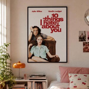May include: A rectangular movie poster for 10 Things I Hate About You featuring Julia Stiles and Heath Ledger. The poster has a white background with red text and shows the two actors sitting in a brown armchair. It is framed in a thin black frame.